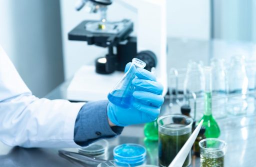A mão de uma pessoa aparece em um laboratório segurando um recipiente com produto químico.