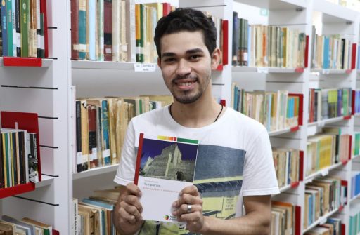 A foto tem como fundo prateleiras de livros da biblioteca da UEMASUL. O historiador Wekslley Machado mostra o livro de sua autoria: Templários: Entre a pureza e a sodomia no século XIV.