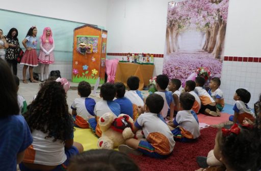 Crianças de escolas municipais reunidas em uma sala e sentadas em um tapete vermelho. A frente temos os acadêmicos vestidos com fantasias de personagens infantis.