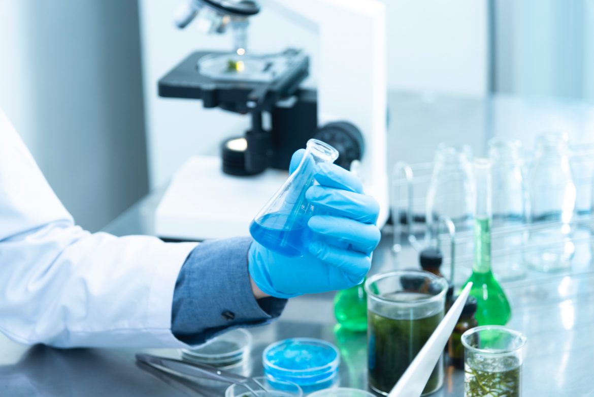 A mão de uma pessoa aparece em um laboratório segurando um recipiente com produto químico.