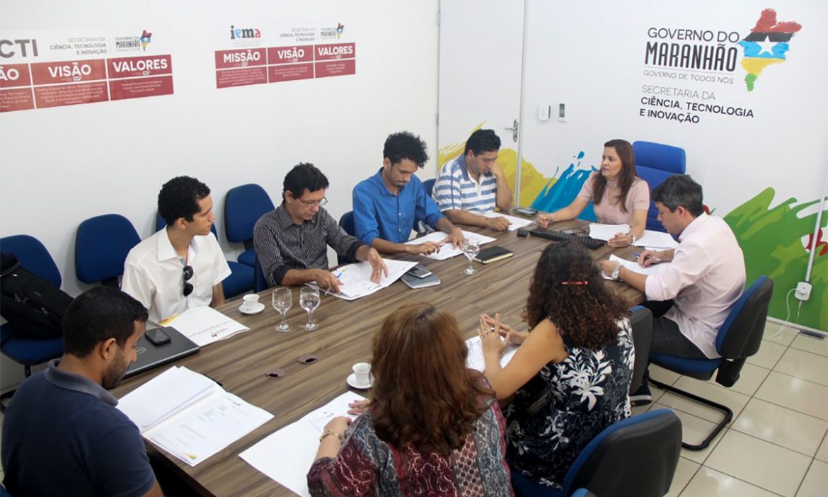 Um grupo de pessoas aparece sentado ao redor de uma mesa em reunião.