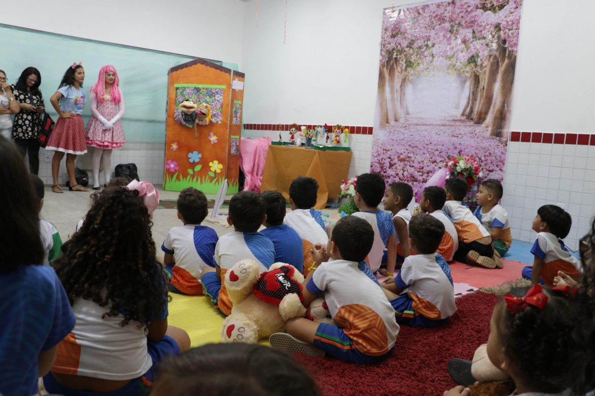 Crianças de escolas municipais reunidas em uma sala e sentadas em um tapete vermelho. A frente temos os acadêmicos vestidos com fantasias de personagens infantis.