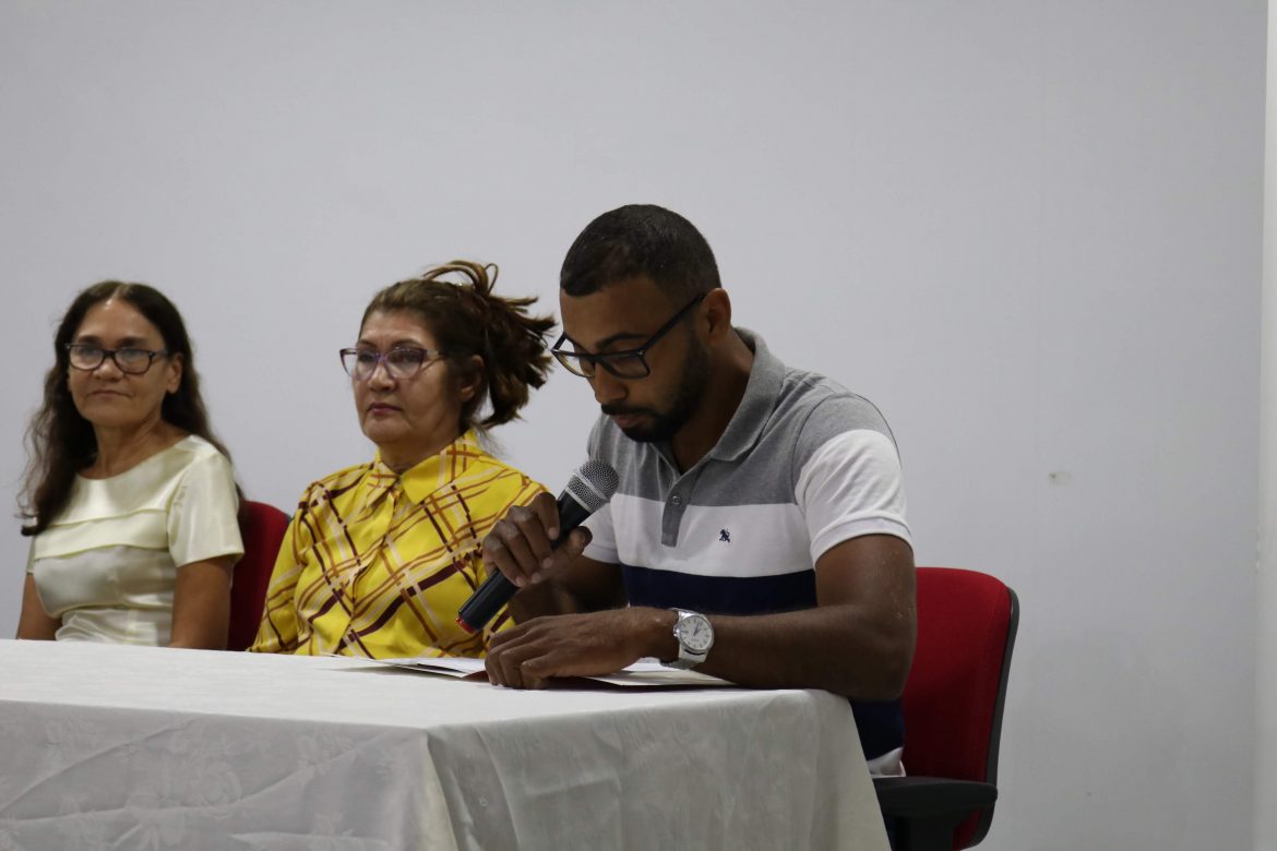 Sentados à mesa do auditório da UEMASUL, à direita duas professoras, à esquerda o acadêmico, com microfone na mão, lendo seu discurso.