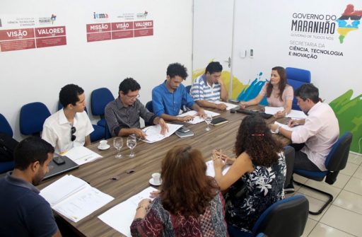 Um grupo de pessoas aparece sentado ao redor de uma mesa em reunião.