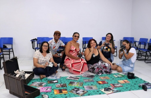 Seis professores sentados no chão, em uma sala de aula, seguram objetos e livros usados na formação.