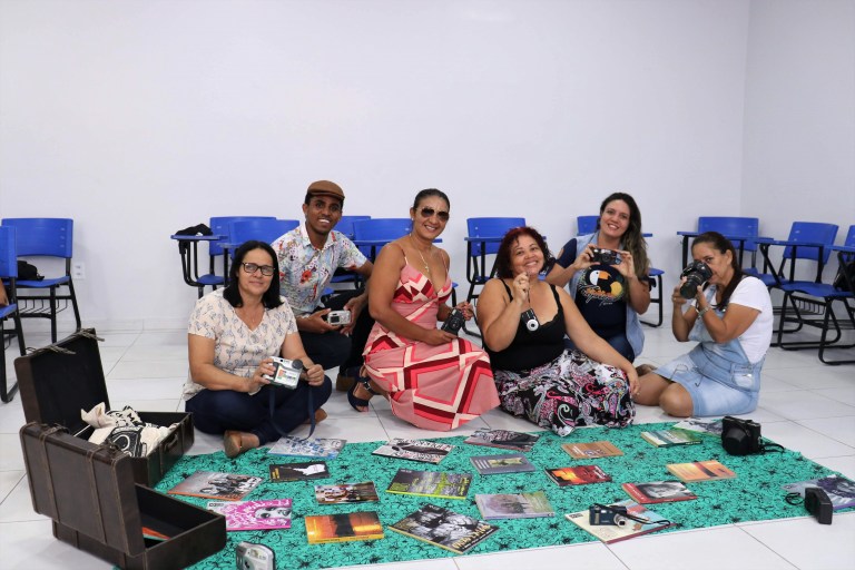 Seis professores sentados no chão, em uma sala de aula, seguram objetos e livros usados na formação.